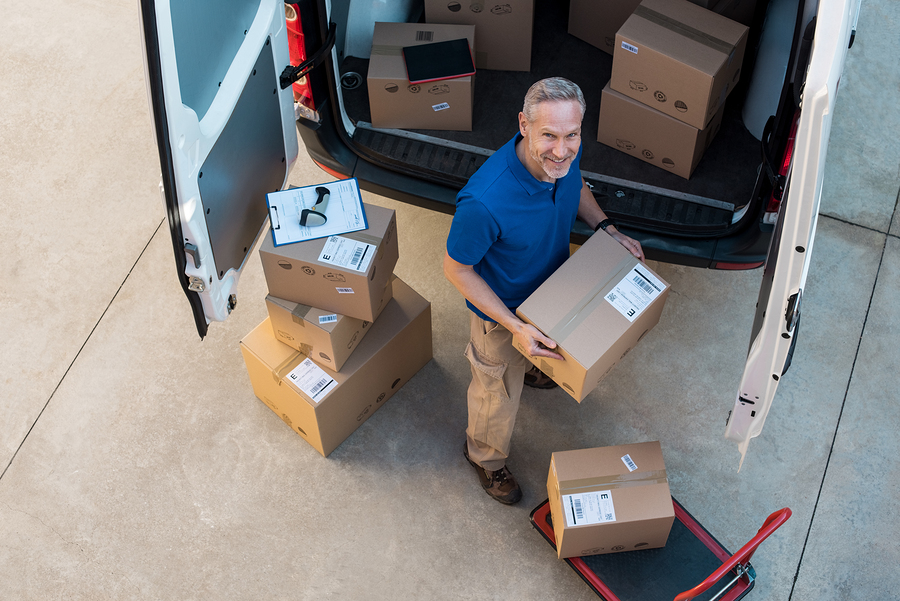 Happy delivery man holding cardboard package and looking at camera. Top view of courier loading packages in van for delivery. Portrait of man working at courier service and carrying parcels on van.